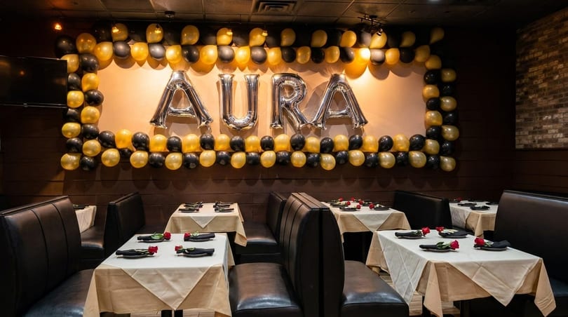 Restaurant interior with black booths, white tablecloths, red roses, and "AURA" balloons on the wall.