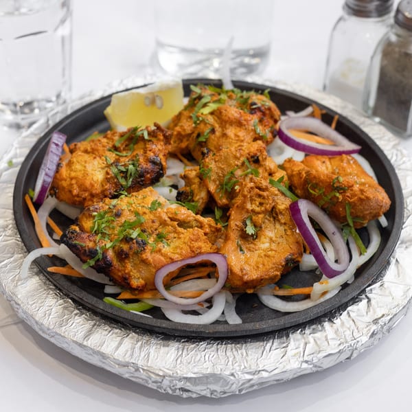 Sizzling chicken tikka with onions, carrots, and lemon on a hot plate, with water and shakers in background.