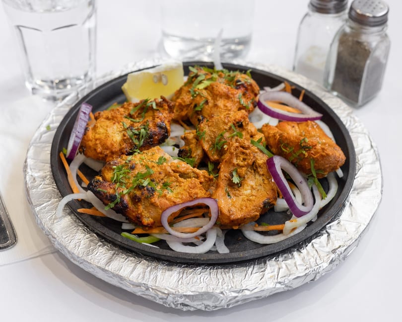 Sizzling chicken tikka with onions, carrots, and lemon on a hot plate, with water and shakers in background.