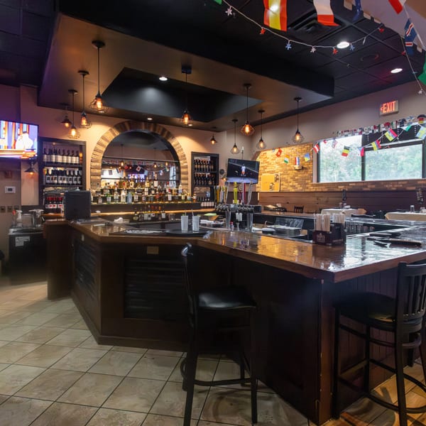Restaurant bar area with dark wood counter, liquor bottles, TVs, and international flags hanging.
