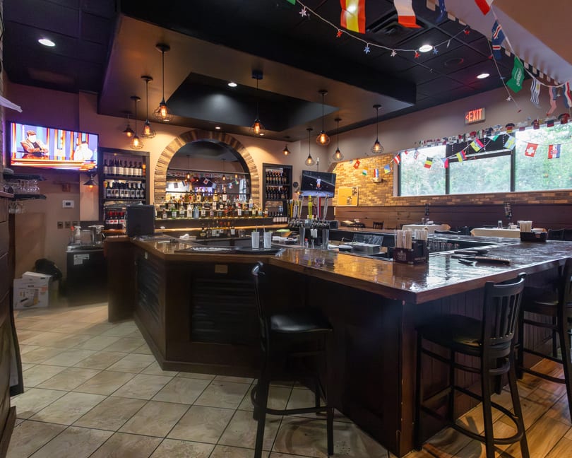Restaurant bar area with dark wood counter, liquor bottles, TVs, and international flags hanging.