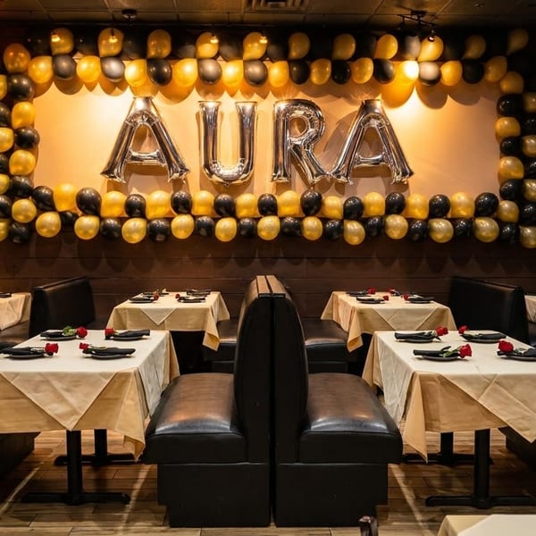 Restaurant dining area with black booths, tables set with white cloths, and a balloon arch spelling "AURA".