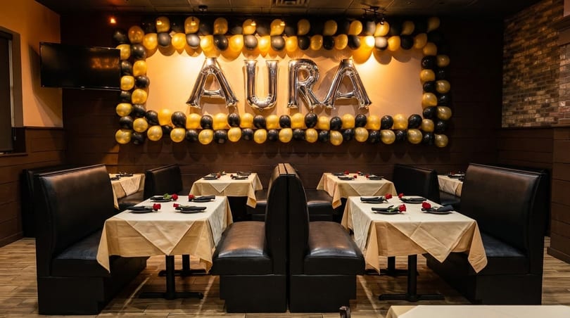 Restaurant dining area with black booths, tables set with white cloths, and a balloon arch spelling "AURA".