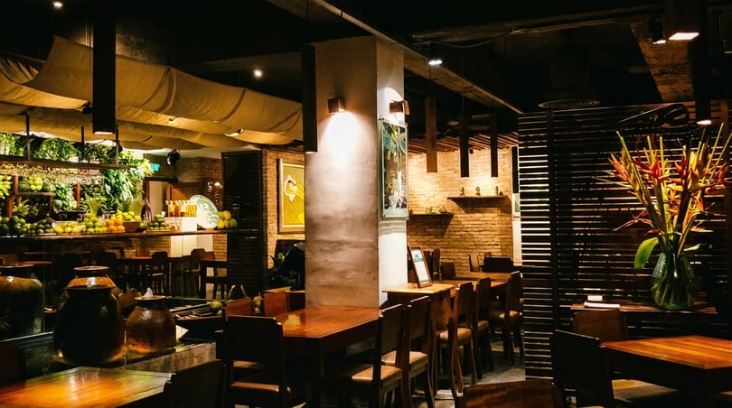 Warmly lit restaurant interior with wooden tables, brick walls, lush greenery, and a fruit display.