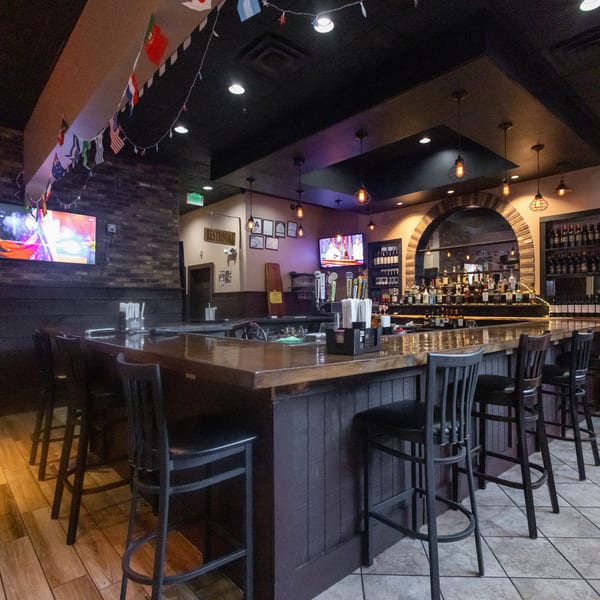 Empty restaurant bar with stools, liquor bottles, multiple TVs, and international flags hanging overhead.