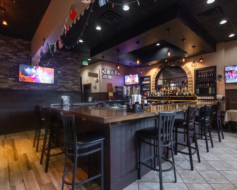 Empty restaurant bar with stools, liquor bottles, multiple TVs, and international flags hanging overhead.